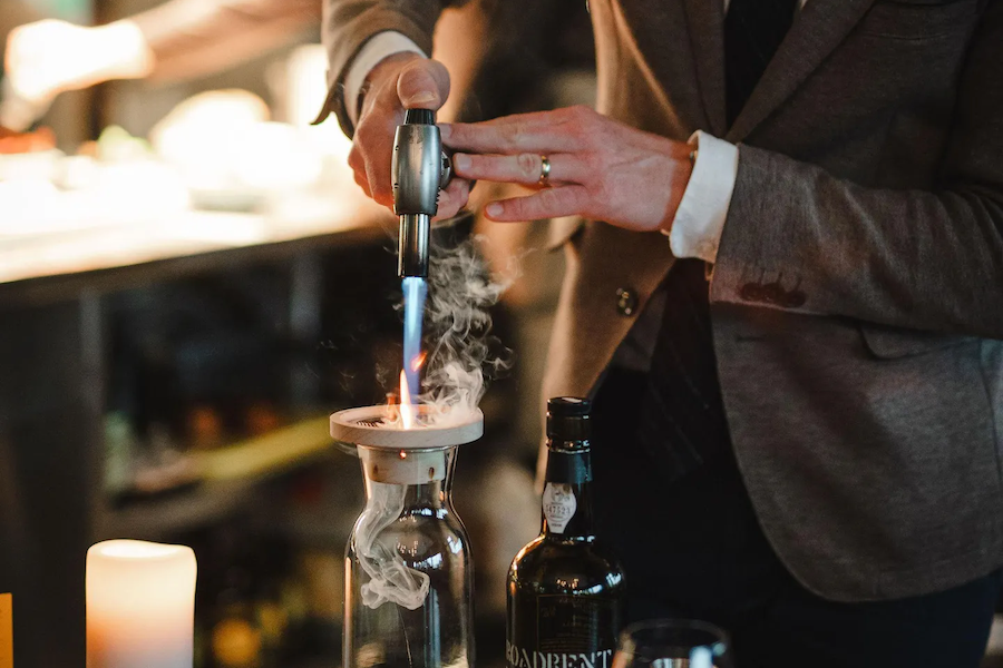A person in a suit uses a torch to create a smoky effect in a glass decanter, with a bottle of liquor nearby.