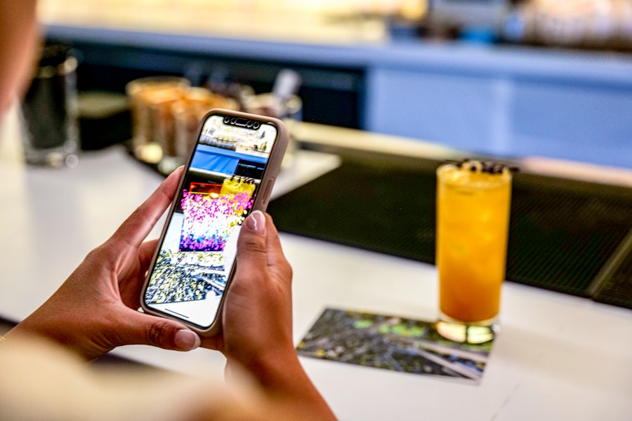 A person holding a smartphone, viewing colorful images transposed onto the camera's image of the tall orange cocktail on the bar counter where they are seated