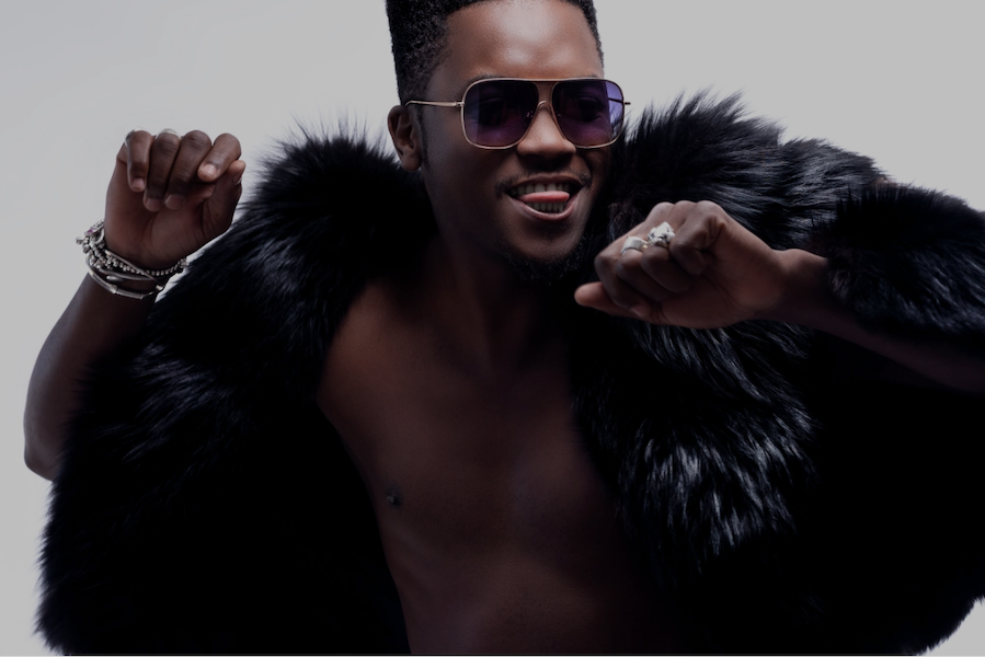 Man wearing sunglasses and a black fur coat, smiling and striking a dance pose.