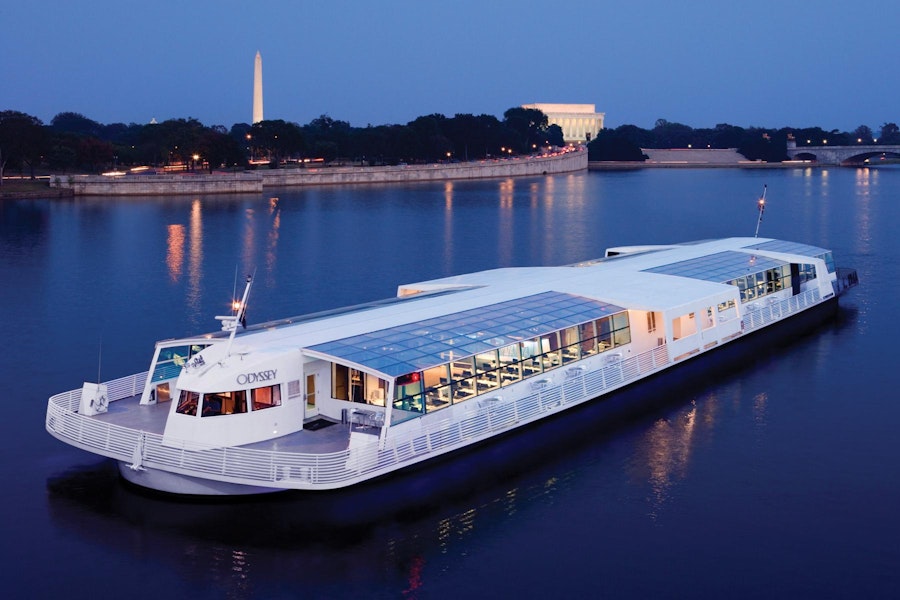A white riverboat named "Odyssey" cruises on calm waters at dusk with the Washington Monument and Lincoln Memorial illuminated in the background.