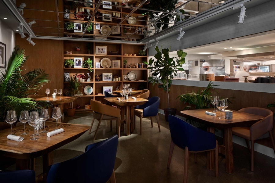 A cozy restaurant interior with wooden tables, elegant table settings, and plush chairs. The space features a wall with decorative shelves, plants, and a large window into the kitchen.