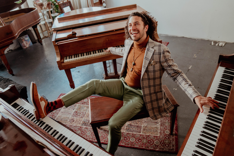Man in a plaid blazer and green pants sitting playfully among several pianos, smiling.