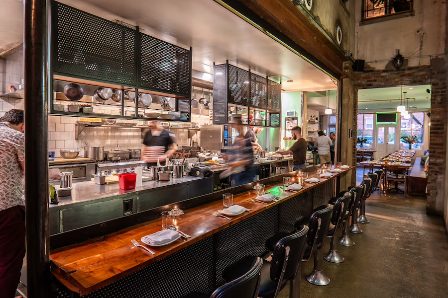 An open kitchen restaurant with a long wooden counter and barstools. Chefs are busy preparing food, and tables are set for diners. The interior features exposed brick and warm lighting.