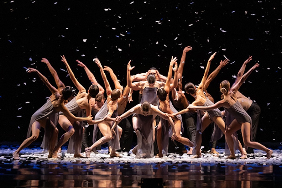 Dancers in earth-toned costumes perform a dramatic ballet scene, surrounding a central figure under a shower of white confetti.