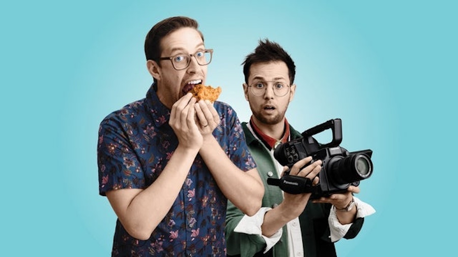 Two men pose against a light blue background. The man on the left, wearing glasses and a patterned shirt, is eating a piece of fried chicken. The man on the right, also wearing glasses and a green and white jacket, is holding a video camera and looking surprised.