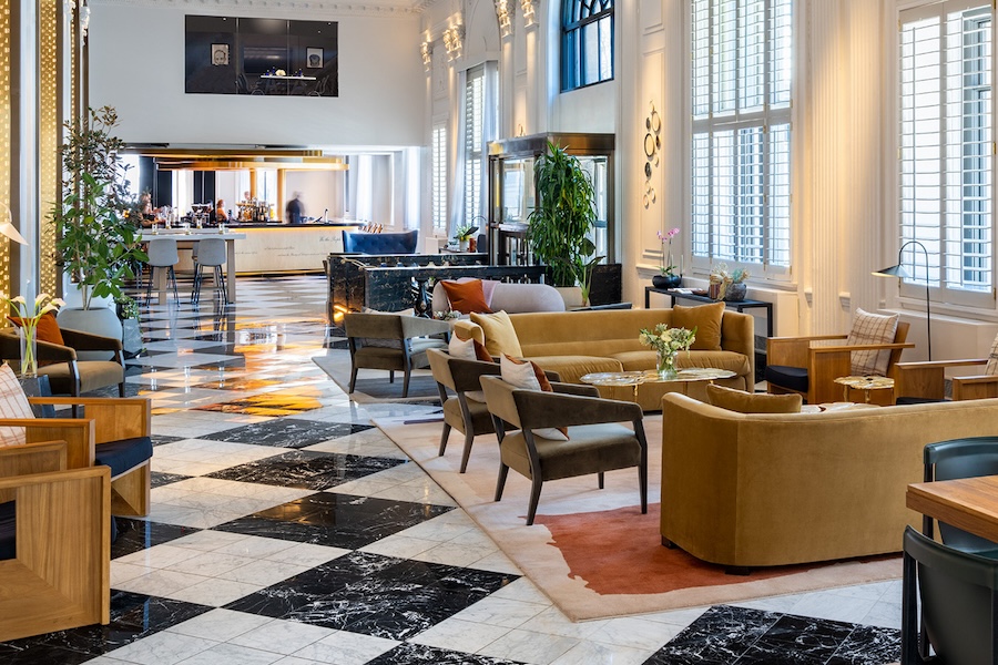 A luxurious hotel lobby with elegant furniture, including sofas, armchairs, and coffee tables, arranged on a polished marble floor. The space is well-lit with large windows and features a bar area in the background.