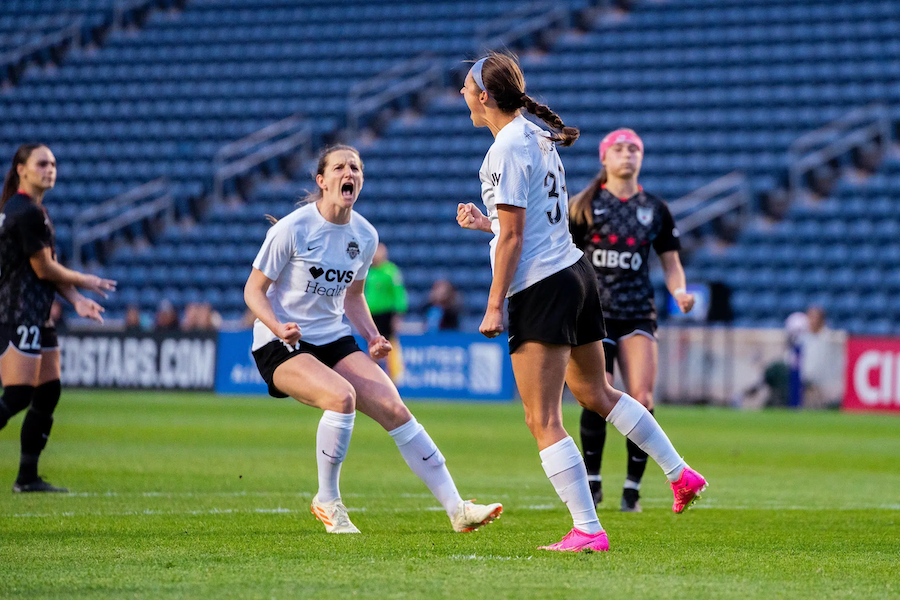 Two Washington Spirit players in white jerseys celebrating passionately on the field during a match, with other players and the stands visible in the background.
