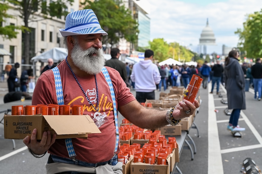 A man with a white beard and a blue checkered hat and suspenders, handing out orange beer cans at an Oktoberfest event in Washington D.C., with the U.S. Capitol building visible in the background.