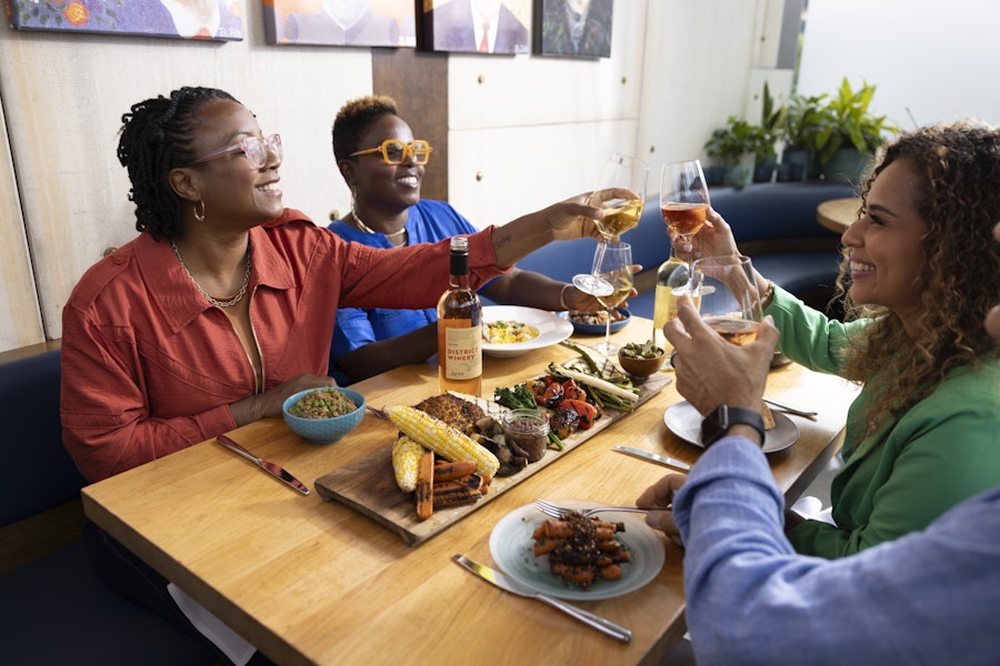 Four people sit around a wooden table in a restaurant, toasting with glasses of wine. The table is set with a variety of dishes, including grilled corn, vegetables, and a bottle of District Winery wine. The atmosphere is lively and the participants appear to be enjoying their meal and company.