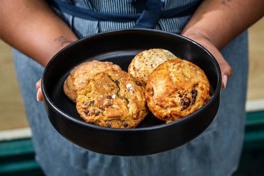 A person holding a black bowl filled with freshly baked cookies and scones, wearing a striped apron.