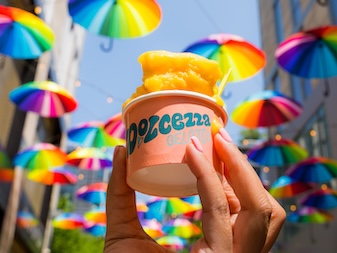 A hand holding a cup of orange gelato with vibrant, rainbow-colored umbrellas hanging overhead in the background.