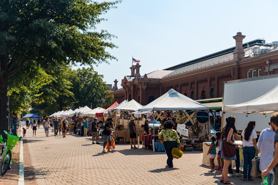 A bustling outdoor market with rows of vendor tents offering various goods. Shoppers walk along the brick-paved path, with the historic Eastern Market building in the background.