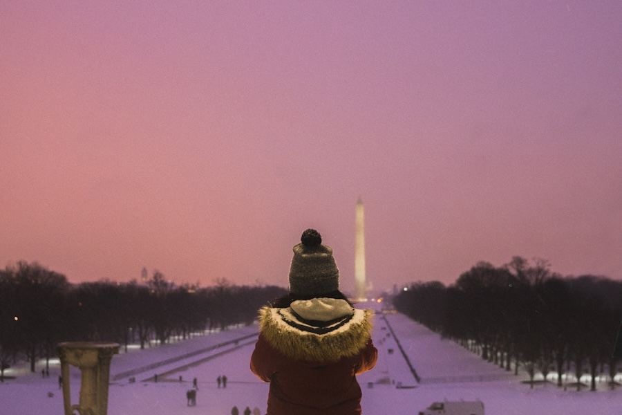 A person in a winter coat and hat stands at the Lincoln Memorial, looking out at the Washington Monument in the distance. The snowy landscape is illuminated by a soft, pinkish sky during twilight.