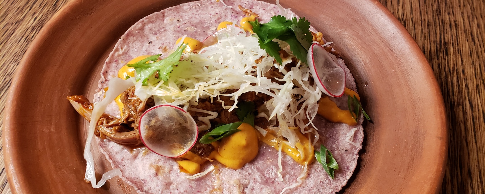 A taco served on a purple corn tortilla, topped with shredded meat, cabbage, radish slices, and fresh cilantro, with drizzles of a creamy orange sauce. The dish is presented on a rustic clay plate.