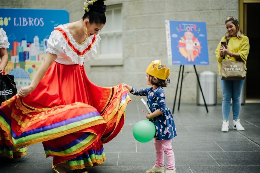 A woman in a colorful traditional dress interacts with a young girl wearing a paper crown and holding a green balloon. The woman holds out her dress, and the girl gently touches it while an onlooker takes a photo in the background.