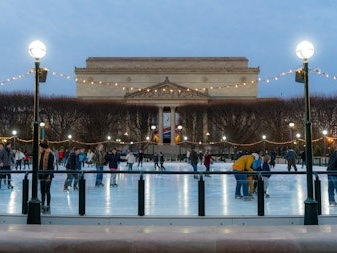 People ice skating at the National Gallery of Art Sculpture Garden rink during the evening, with string lights overhead and the museum building in the background.