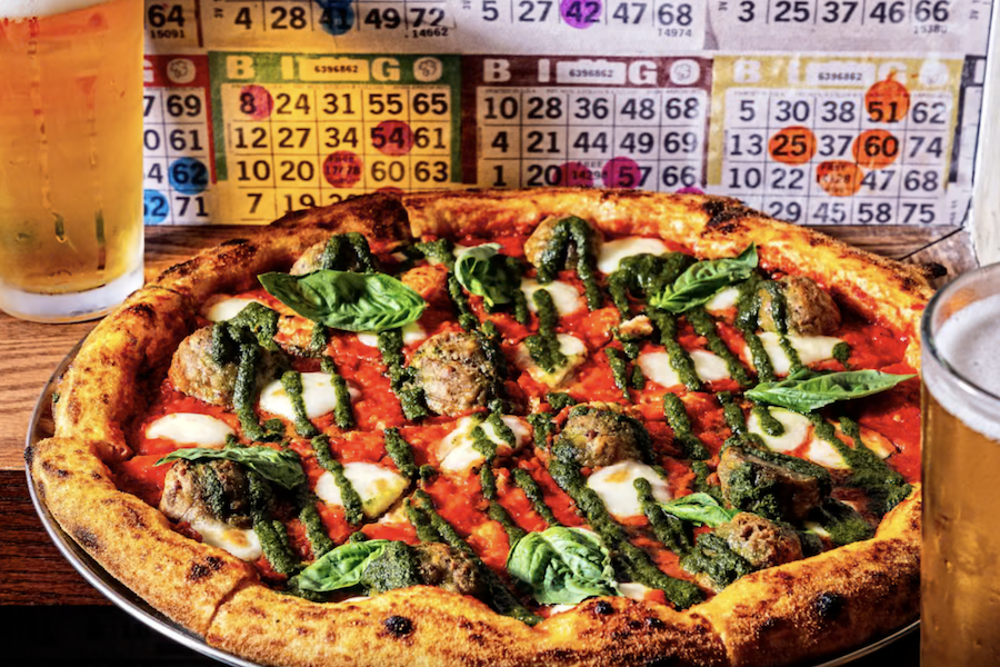 A close-up of a freshly baked pizza topped with meatballs, mozzarella cheese, basil leaves, and pesto drizzle. Two pints of beer are visible next to the pizza, and a colorful bingo card background adds a playful atmosphere to the scene.