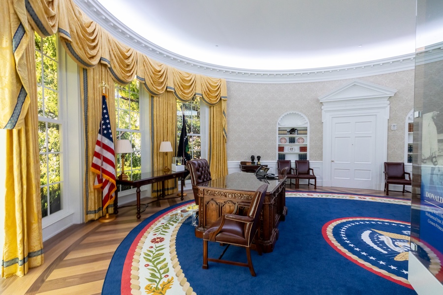 A view of the People's House replica of the Oval Office, showcasing the iconic Resolute Desk, gold curtains, and presidential seal on the carpet, with the American flag and windows providing a bright backdrop.