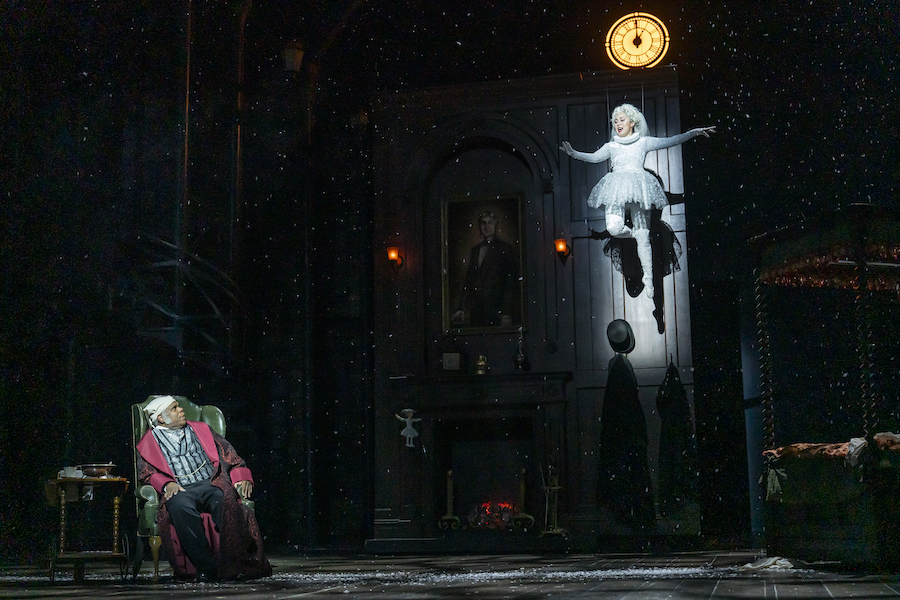  A theatrical scene from "A Christmas Carol" featuring Scrooge in a chair, looking up in surprise at the Ghost of Christmas Past, who appears to float in mid-air amidst a snowy backdrop. A vintage clock and dark, shadowy set add to the dramatic atmosphere.