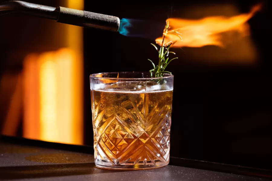 A close-up of a glass filled with a cocktail, topped with a sprig of rosemary being set on fire with a torch, creating a dramatic visual effect.