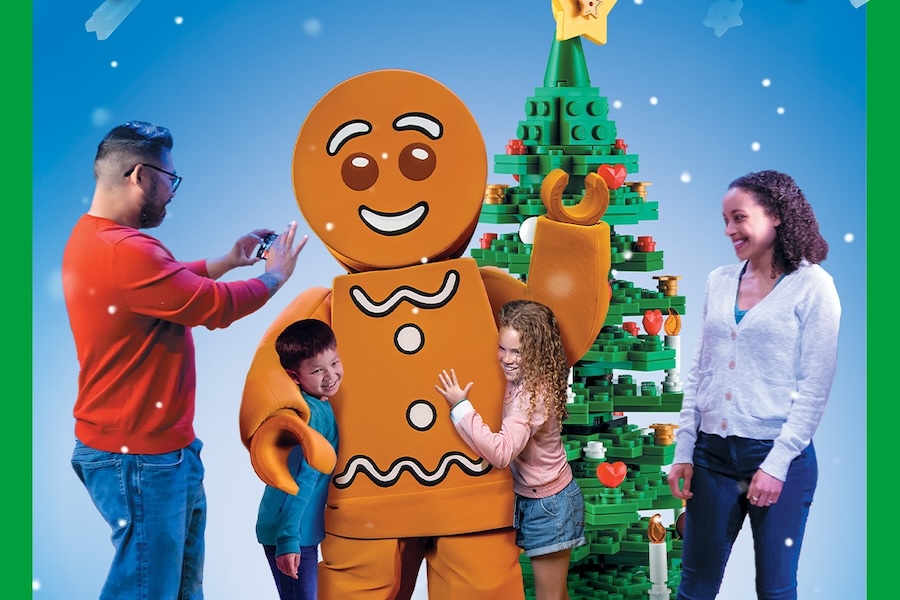 Family posing with a giant gingerbread mascot and a LEGO tree at a holiday event.