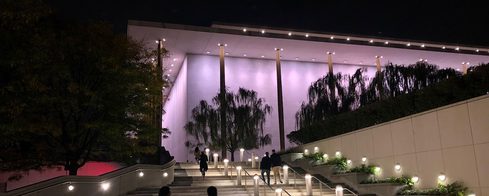 Nighttime view of the Kennedy Center's outdoor stairs, with people walking up and the building illuminated in soft purple lights.