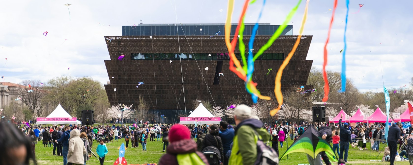 A colorful scene from the Blossom Kite Festival with kites flying in the sky in front of the National Museum of African American History and Culture.