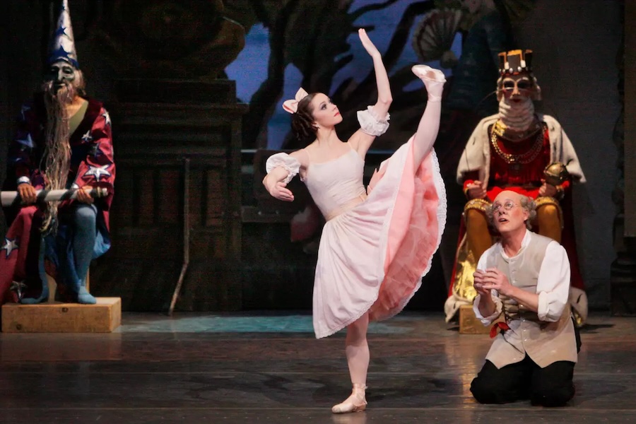 A ballet dancer performs a high kick on stage beside a kneeling man, with theatrical figures in the background.