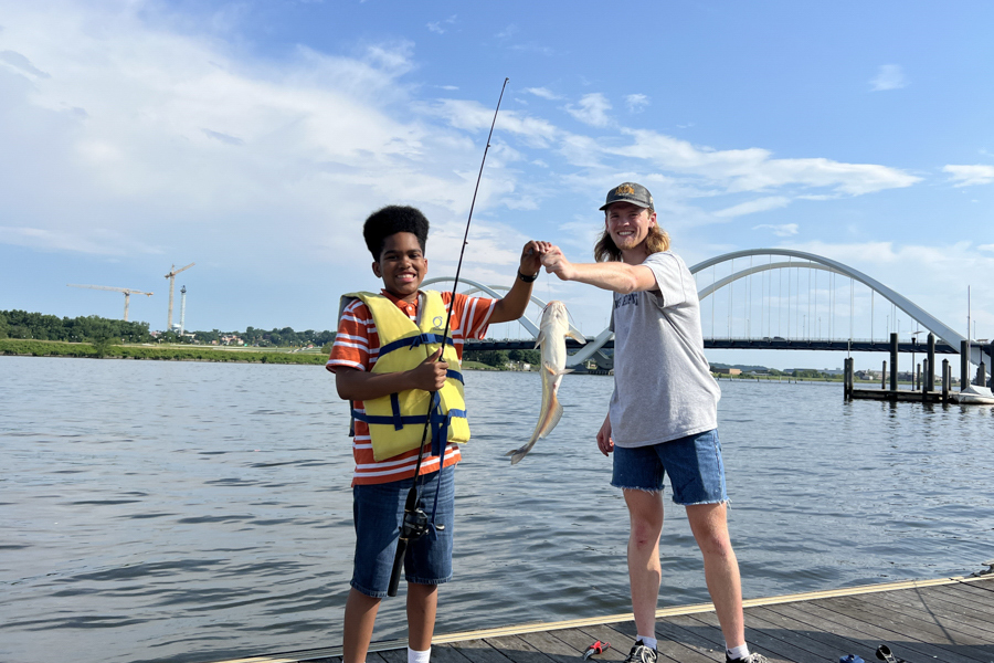 A smiling boy wearing a life jacket holds a fishing rod while shaking hands with an older person holding a freshly caught fish, standing on a dock with a bridge and water in the background.