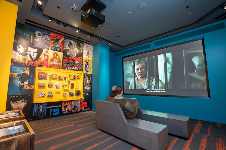 Visitors sit on modern benches watching a spy film at an exhibit featuring a large screen, vibrant blue walls and a collage of classic espionage movie posters.