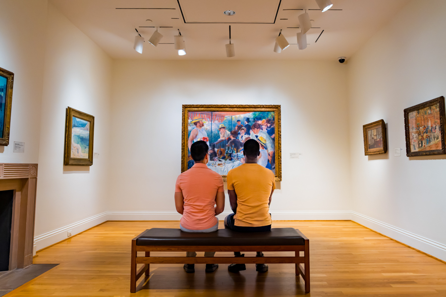 Two people in orange shirts sit on a bench, admiring an Impressionist painting in a quiet gallery.