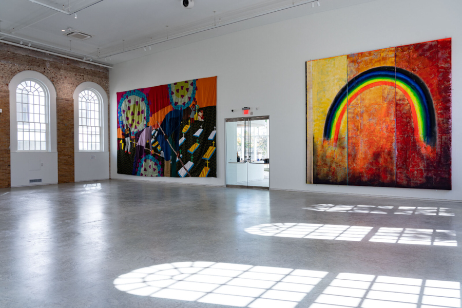 A bright, modern gallery space features large contemporary artworks on white walls, with sunlight casting geometric shadows on the polished floor.