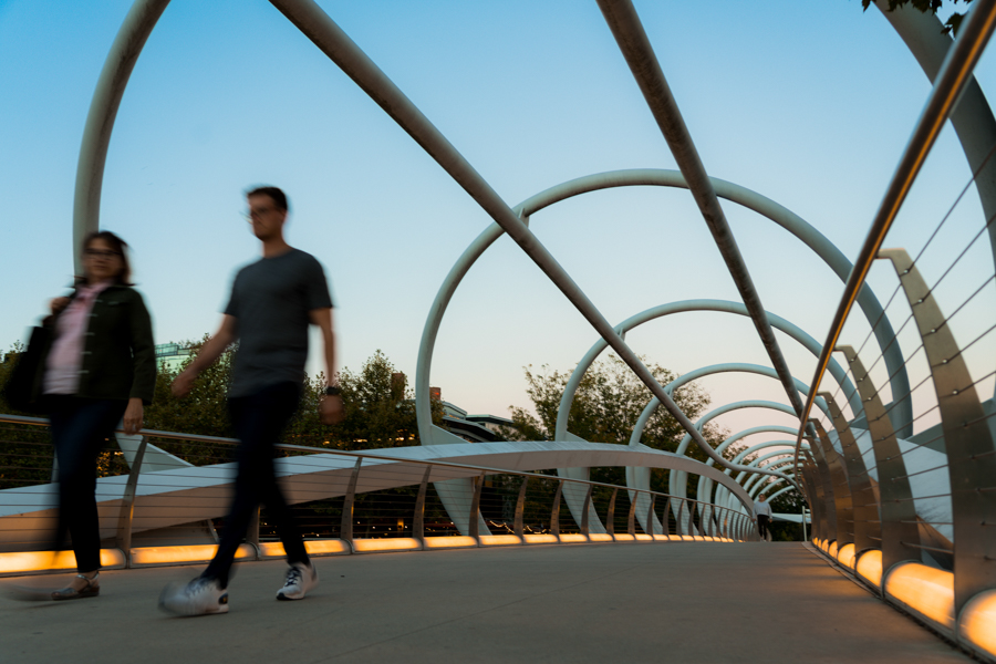 Two people walk across the illuminated Yards Park Bridge at dusk, surrounded by its signature curved metal arches.