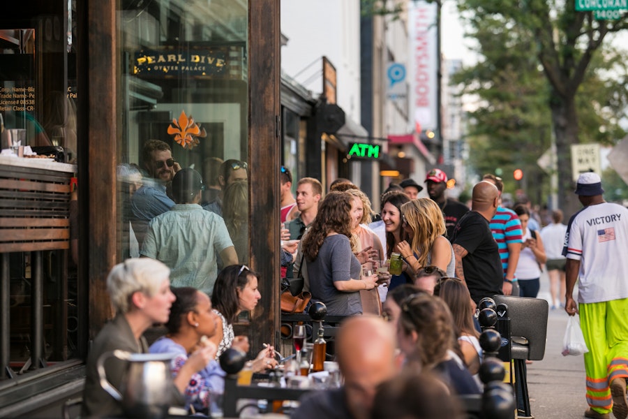 Eine lebhafte Menschenmenge versammelt sich vor dem Pearl Dive Oyster Palace, unterhält sich und genießt Getränke auf dem belebten Bürgersteig der 14th Street.