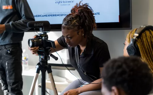 A young Black woman prepares a camera to record video and sound. She is wearing her Academy polo that says "Coolidge Media"
