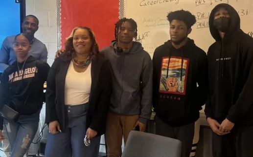 A Black woman professional smiles and stands with a group of Black high school students at Maya Angelou Public Charter School.
