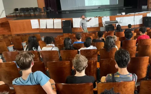 An educator stands in front of students seated in the auditorium at Jackson-Reed High School
