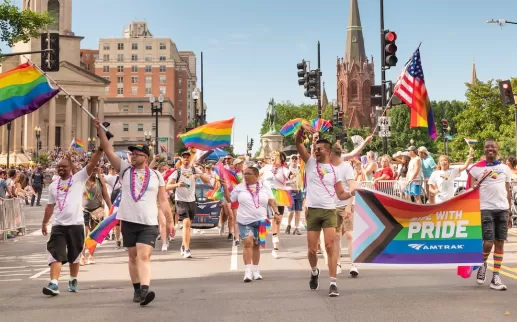 A vibrant group marches down a city street holding rainbow flags and a “Ride With Pride” banner during a Pride Parade, cheered on by festive crowds.