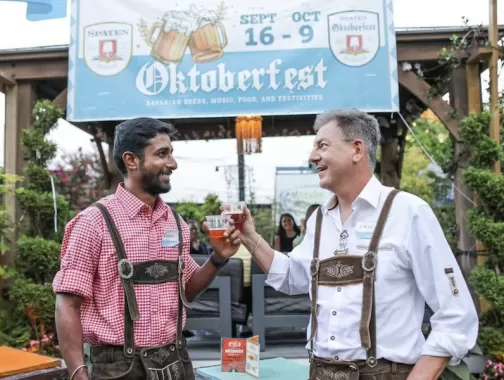 Two men in traditional Oktoberfest attire clinking glasses of beer at an outdoor Oktoberfest event.