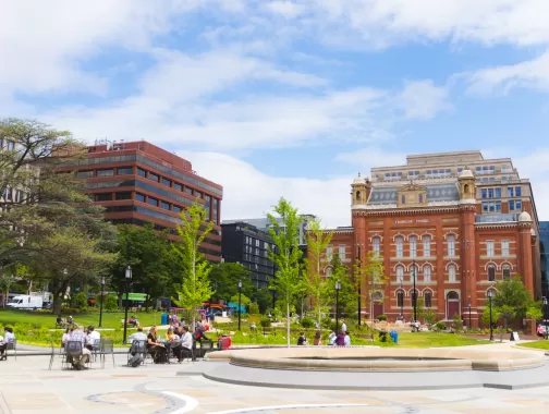 A sunny day in Franklin Square, Washington D.C., with people enjoying the park near the historic red-brick Franklin School building and surrounding office buildings.