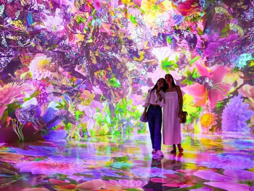 Two visitors stand in a vibrant, immersive digital art installation at ARTECHOUSE, surrounded by colorful, glowing flowers and butterflies.