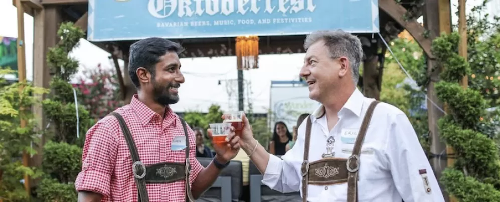 Two men in traditional Oktoberfest attire clinking glasses of beer at an outdoor Oktoberfest event.