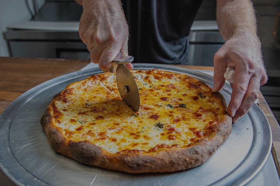Close-up of a person using a pizza cutter to slice a freshly baked cheese pizza on a metal tray.