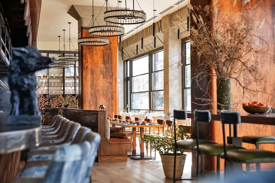 Stylish restaurant interior with rustic decor, large windows, and wooden furnishings. Bar stools line the counter, and tables are set for dining. Chandeliers and plants add to the warm atmosphere.