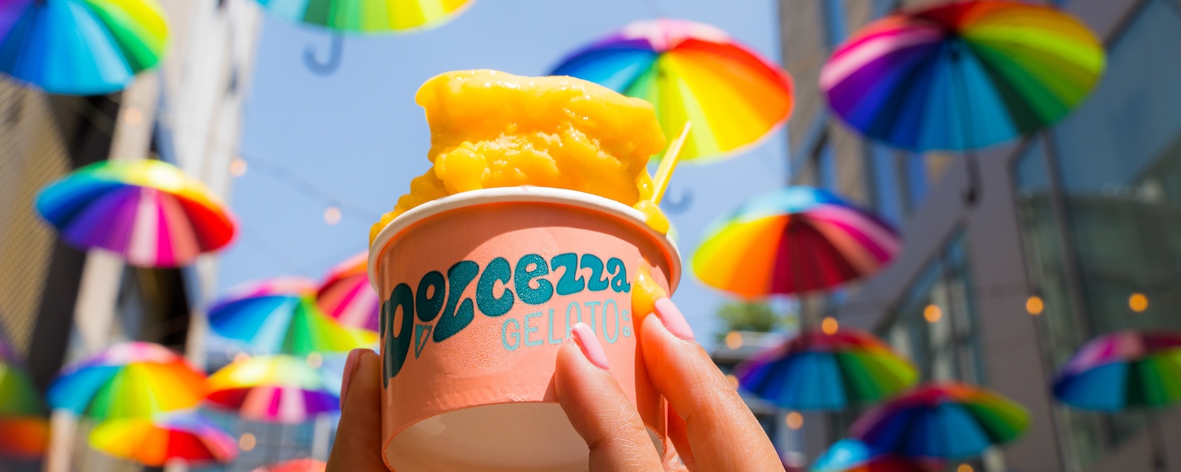 A hand holding a cup of orange gelato with vibrant, rainbow-colored umbrellas hanging overhead in the background.