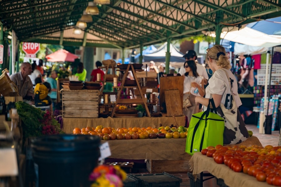  A shopper browses fresh produce at a vibrant outdoor market, with tables full of tomatoes and other goods under a green-roofed pavilion.