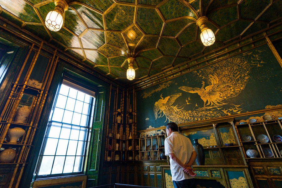 A visitor admires the intricate golden artwork of birds on the wall of a richly decorated room with a green and gold ceiling in an Asian art museum.