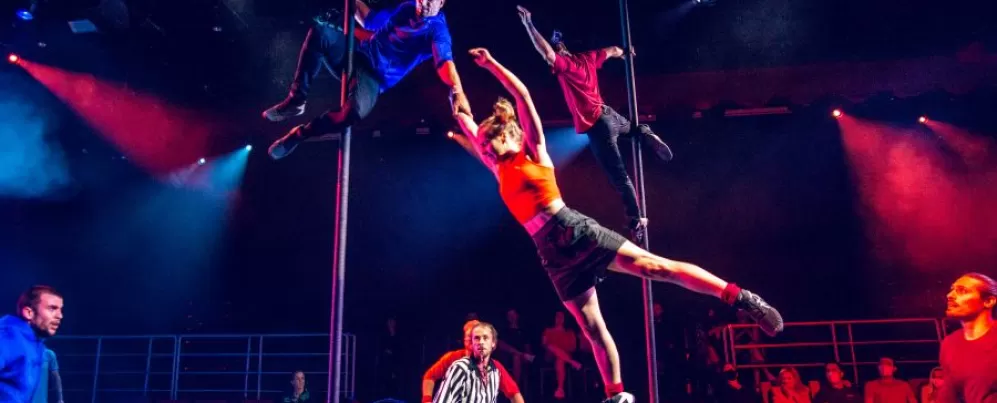 Acrobats perform daring moves on poles while a referee and audience watch during a high-energy scene from Duel Reality.